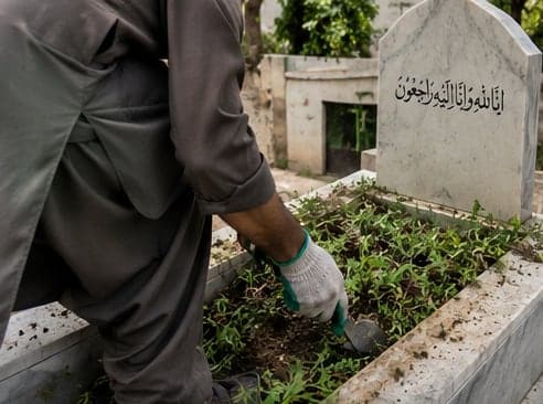 Gloved hands clearing weeds from a grave in a Lahore graveyard
